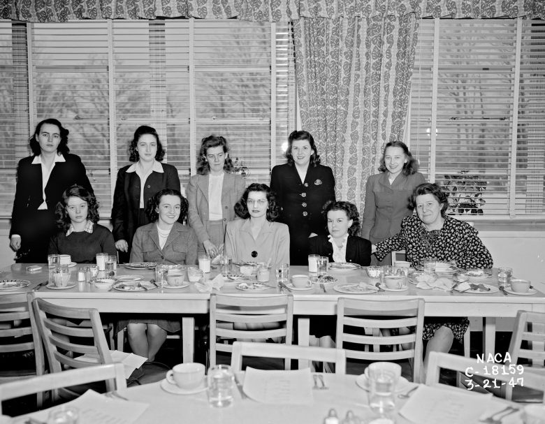 A black and white photo of ten women, with five standing behind five seated at a table.