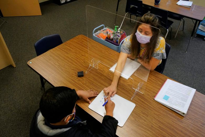 A teacher and student are separated by plexiglass as they sit across from each other at a desk.