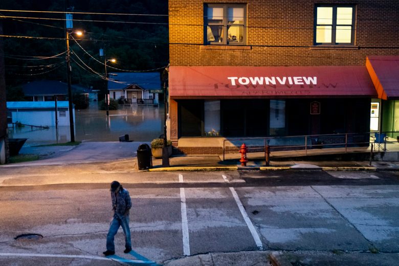 A man walks from a store with lighted rooms above it. In the background, homes are flooded.