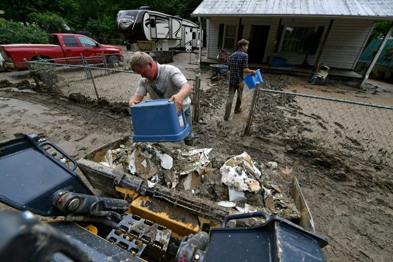 Two men dump buckets of ruined wallboard removed from a home. The yard they are walking through is filled with mud.