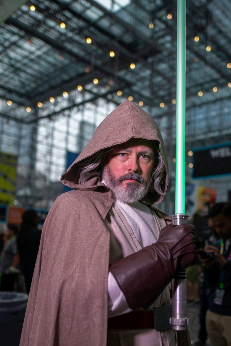 A man in a hooded brown cloak and dark leather gloves stands inside a tall atrium, holding a blue-green pole.