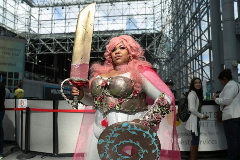 A woman holds a large sword as she poses in a pink wig, chest armor, a shield and a white-and-pink dress.