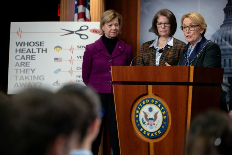 Three women wearing formal blazers stand at a wooden podium, next to a sign that says 'Whose health care are they taking away?'