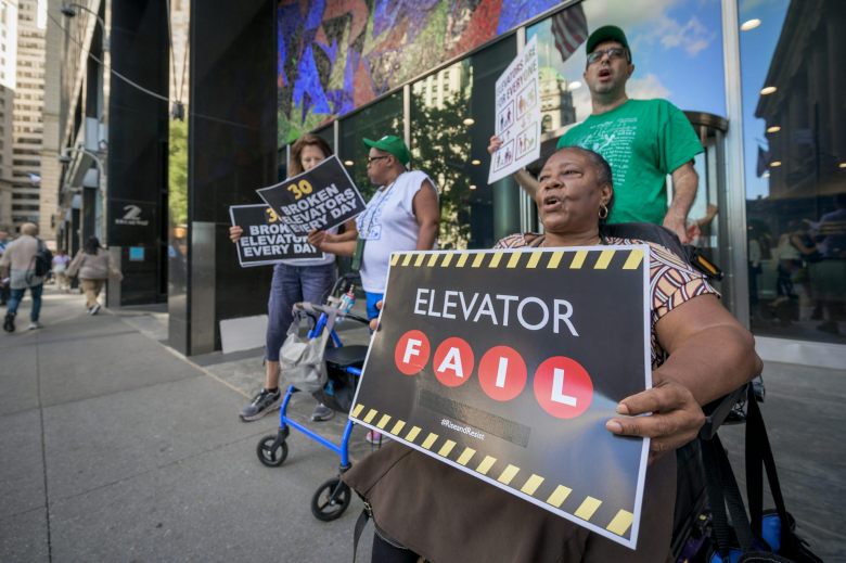 A group of people stand and sit, in a wheelchair, on a street. They hold black and white signs. One of them says 'Elevator Fail.'